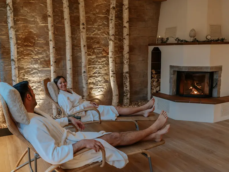 Ruhebereich vor dem Kamin - © Wellness-Ferienhof Lang ein Paar liegt im Bademantel auf Liegen vor dem Kamin