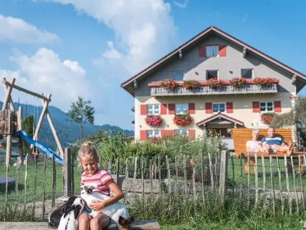Ferienhof Felder - Urlaub auf dem Bauernhof © www.ferienhofffelder.de Mädchen sitzt im Garten mit einer Ziege. Im Hintergrund ein Bauernhof mit Spielplatz und den Eltern im Liegestuhl